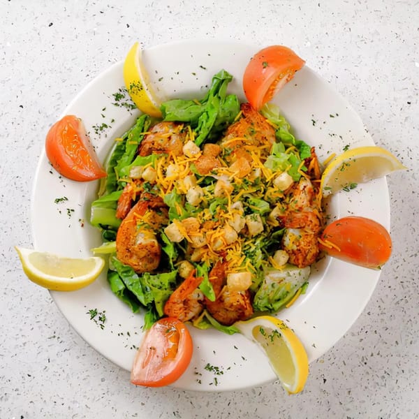 A vibrant shrimp salad with lettuce, cheese, croutons, and lemon and tomato wedges on a white plate.